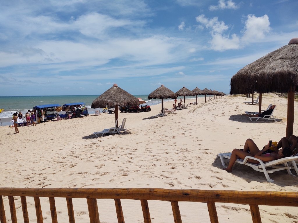 #PraCegoVer Audiodescrição: fotografia de praia com várias barracas de palhas com espreguiçadeiras, com céu azul e uma cerca em primeiro plano