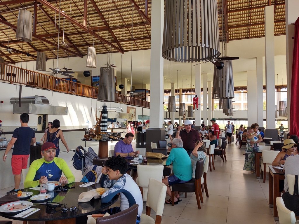 #PraCegoVer Audiodescrição: fotografia interna de restaurante amplo, com várias mesas e teto alto. Muitas pessoas circulando e sentadas