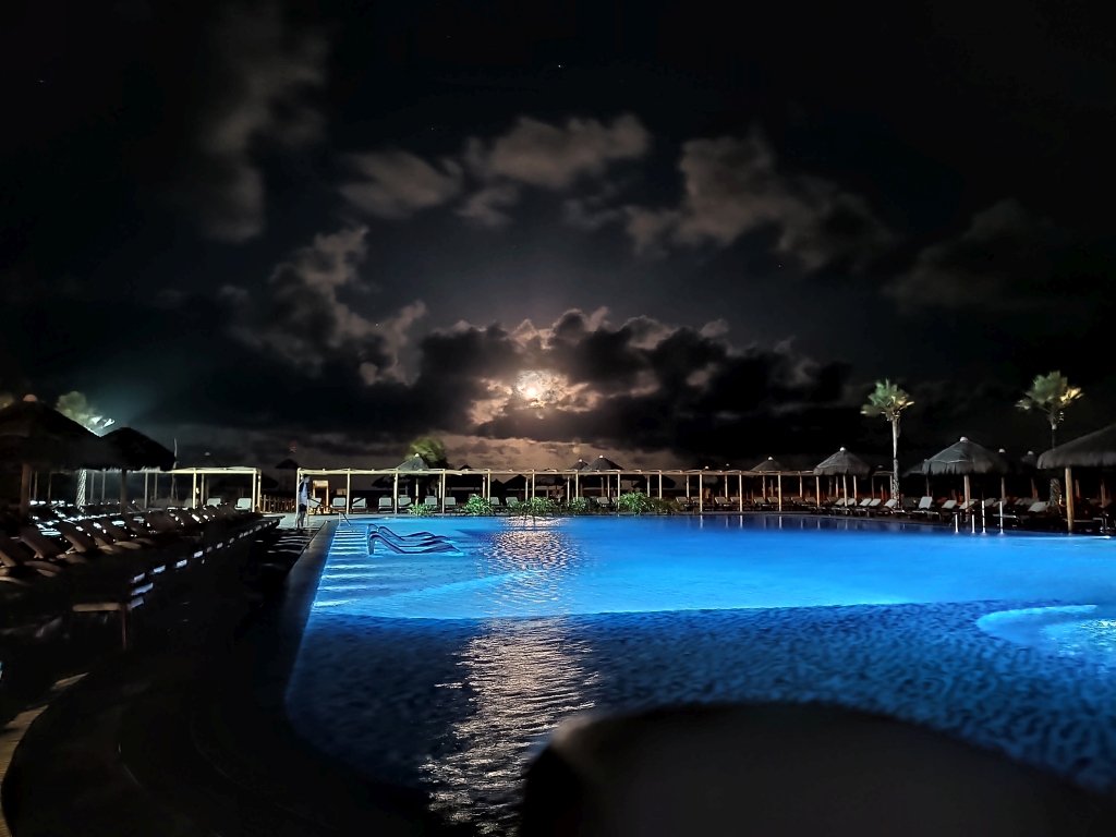 #PraCegoVer Audiodescrição: fotografia noturna com uma piscina iluminada de azul em primeiro plano, uma lua semi encoberta por nuvens