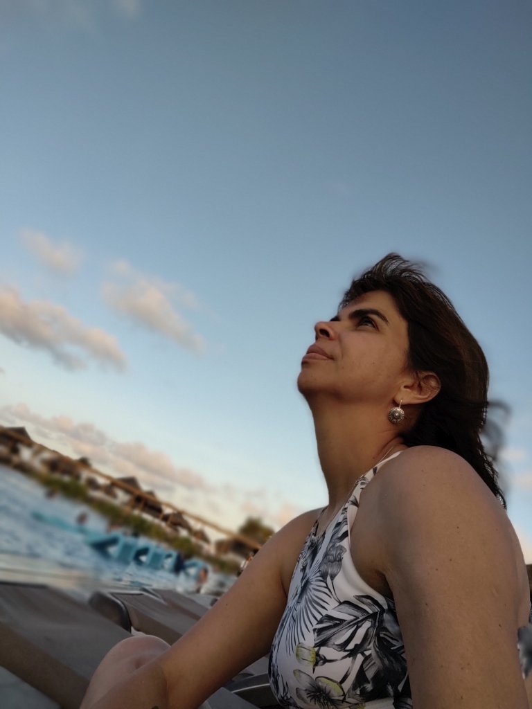 #PraCegoVer fotografia com mulher de perfil olhando para cima em primeiro plano na parte inferior da imagem, com fundo de piscina e céu azul desfocado