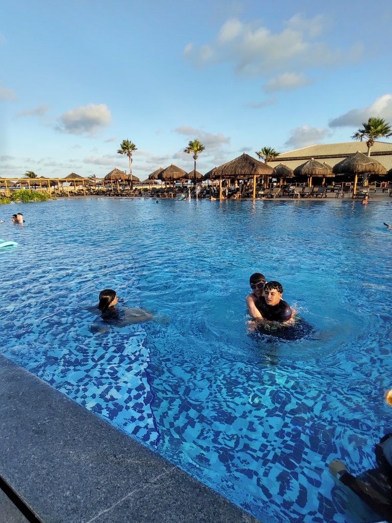 #PraCegoVer Fotografia vertical de piscina azul com três pessoas dentro dela, ao fundo coqueiros, céu de entardecer com algumas nuvens