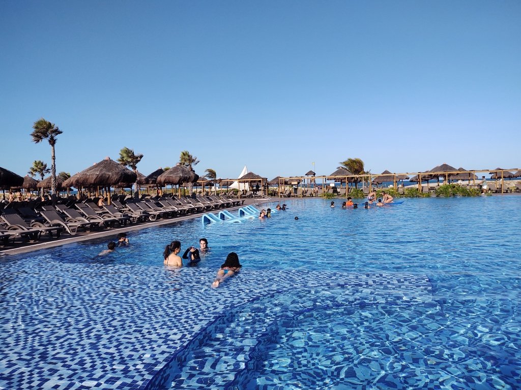 #PraCegoVer Fotografia com grande piscina azul rodeada por espreguiçadeiras e coqueiros. Praia ao fundo. Pessoas na piscina conversando. Céu azul