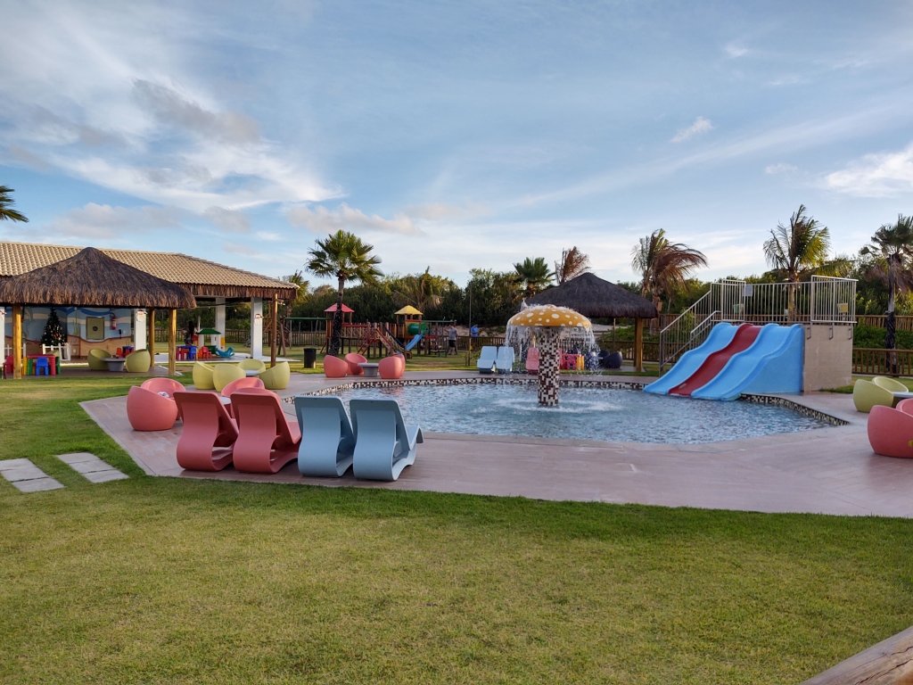 #PraCegoVer Audiodescrição: foto de piscina infantl, com espreguiçadeiras coloridas na frente e tobogãs. Céu azul com algumas nuvens