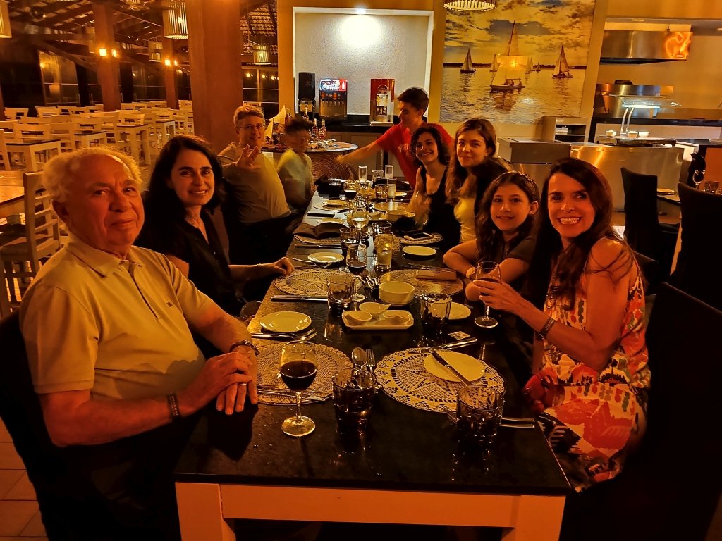 #PraCegoVer Audiodescrição: fotografia noturna de mesa em restaurante com 9 pessoas olhando para a foto