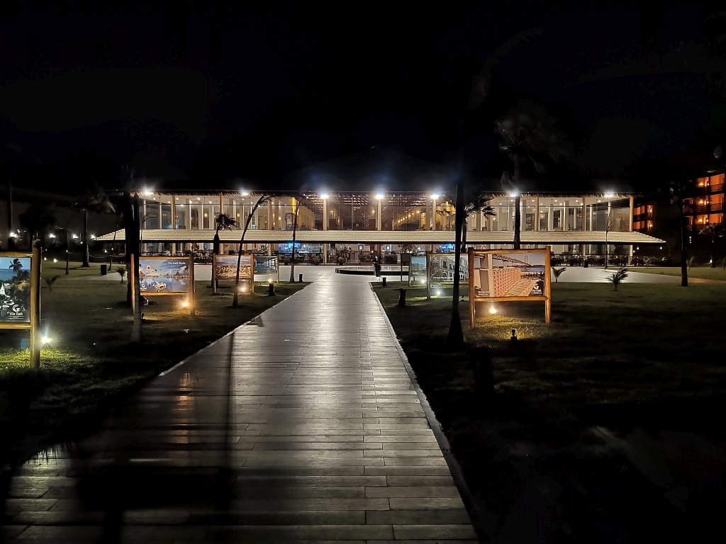 #PraCegoVer Audiodescrição: fotografia noturna de um prédio iluminado com alguns painéis na frente e uma passarela em primeiro plano