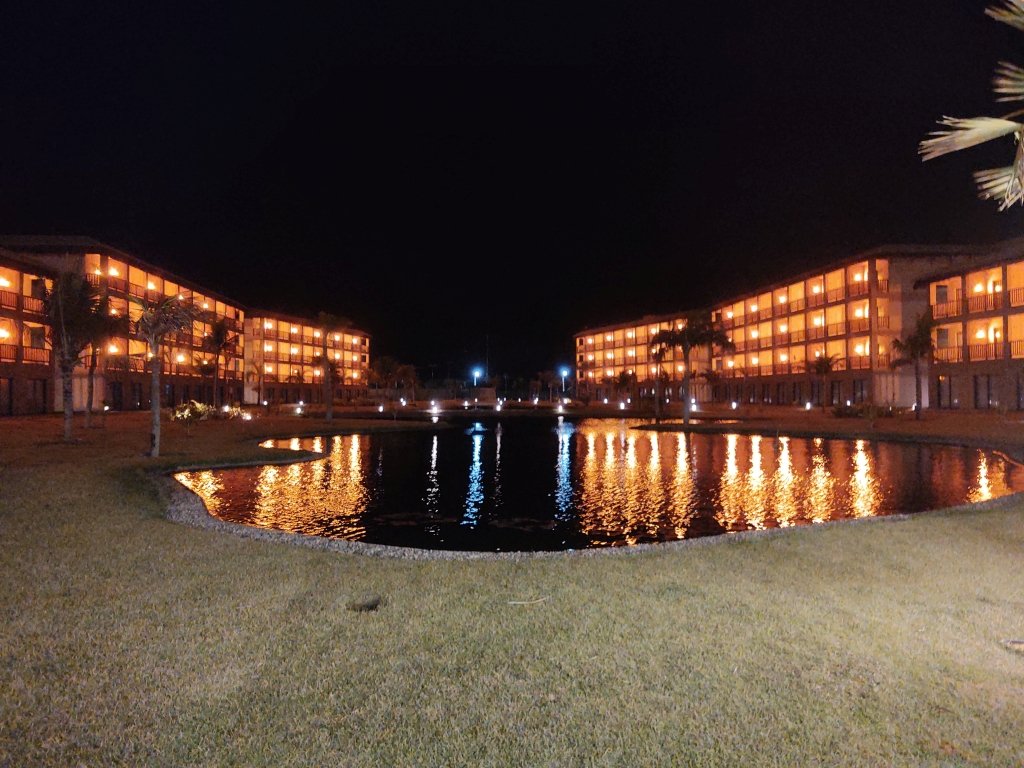 #PraCegoVer Audiodescrição: fotografia noturna de dois prédois de 4 andares iluminados, tendo à frente um lago e no primeiro plano um gramado